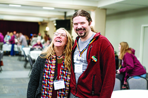 A man and a woman stand close together in a dining hall, smiling and laughing during a casual moment.