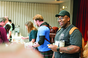 A smiling man in a black shirt, glasses, and a baseball cap holds a drink and food plate while standing in a buffet line.
