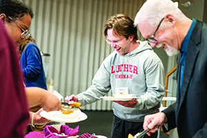 A young man wearing a grey Luther Seminary sweatshirt laughs while filling a plate at a buffet alongside other attendees.