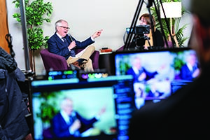 A man in a suit speaks into a microphone during a filmed interview, viewed through a video camera monitor in the foreground.