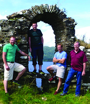 Four adult men smiling and posing together inside and around an ancient, weathered stone archway with a scenic lake and mountains in the background.