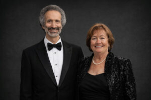 A formal portrait of a man in a black tuxedo and bowtie standing next to a woman in a black sequined jacket and pearl necklace.
